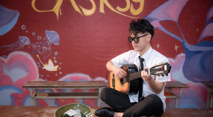 Young musician playing acoustic guitar indoors, busking next to a helmet with money.