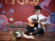 Young musician playing acoustic guitar indoors, busking next to a helmet with money.