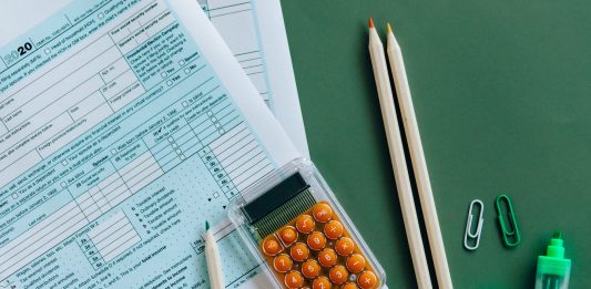 Flat lay of tax forms, calculator, pencils, and clips on green surface, ideal for finance or accounting themes.