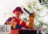 A clown in vibrant costume entertains the crowd with a puppet at an outdoor carnival.