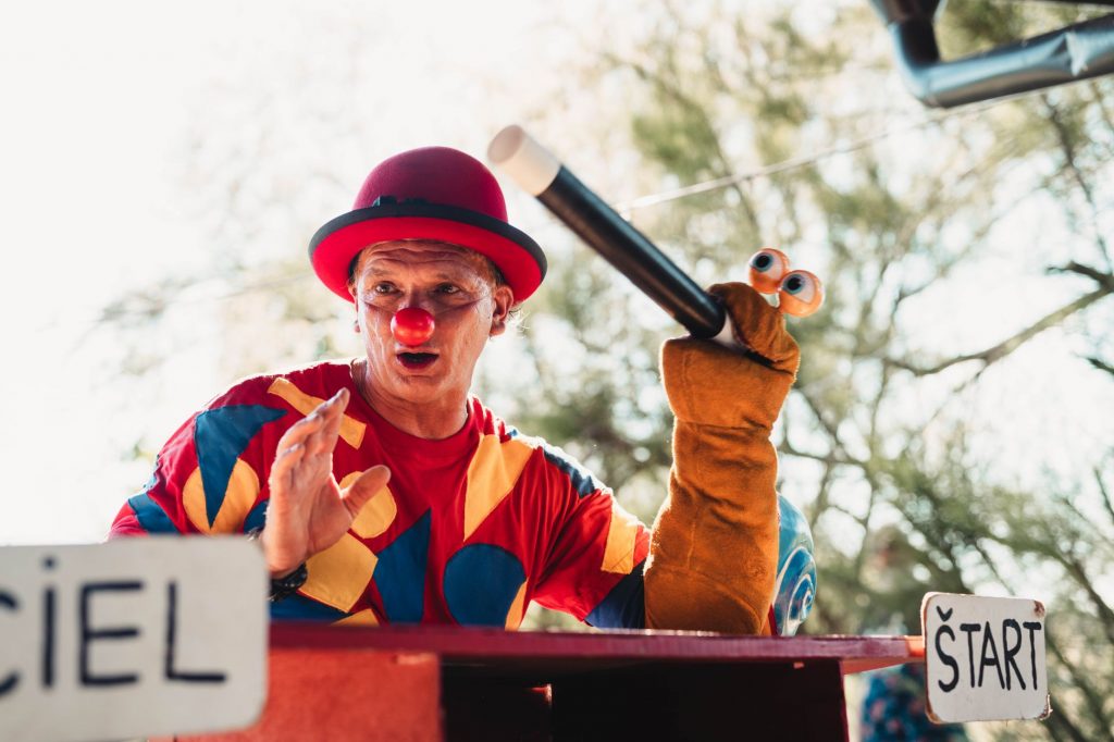 A clown in vibrant costume entertains the crowd with a puppet at an outdoor carnival.