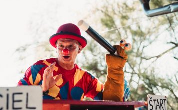 A clown in vibrant costume entertains the crowd with a puppet at an outdoor carnival.