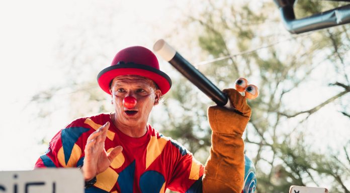 A clown in vibrant costume entertains the crowd with a puppet at an outdoor carnival.