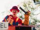 A clown in vibrant costume entertains the crowd with a puppet at an outdoor carnival.