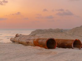 ÚLTIMA HORA: Autoridad de servicios busca más fondos de desarrolladores Rusty pipes at a coastal construction site with scenic sunset view.