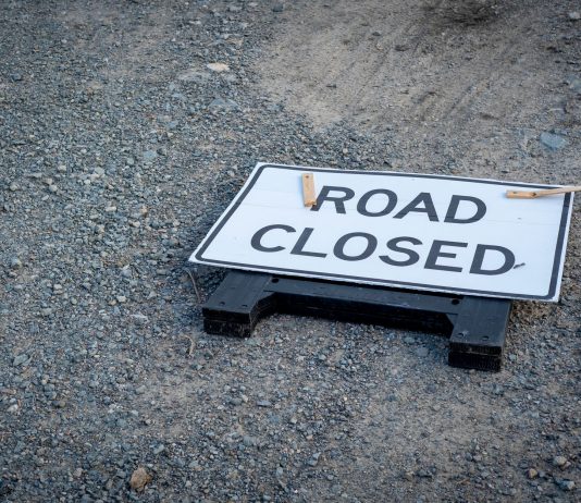 A road closed sign lies on a gravel pathway, indicating a blocked road ahead.