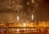 Vibrant fireworks light up the night sky over a bustling waterfront city, captivating an excited crowd below.
