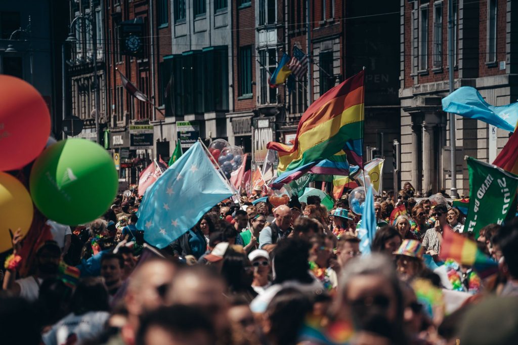 Crowded street filled with people celebrating a lively Pride parade, waving rainbow flags and colorful balloons.