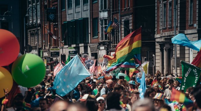 Crowded street filled with people celebrating a lively Pride parade, waving rainbow flags and colorful balloons.