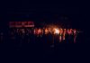 A large group of people gathered around a bonfire at night, featuring a vibrant outdoor atmosphere.