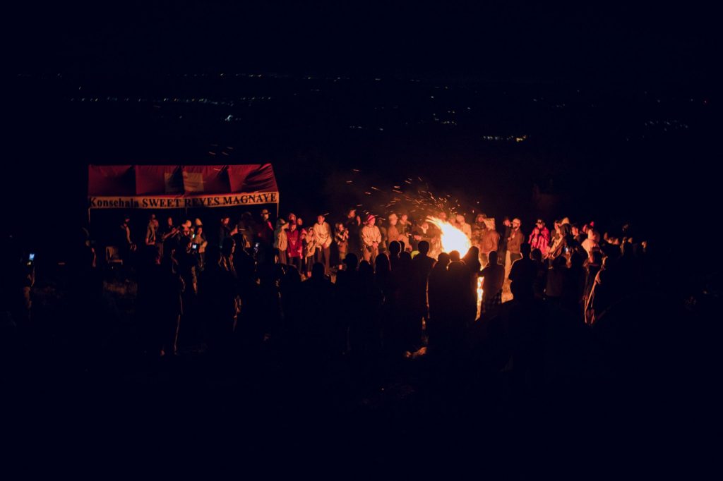 A large group of people gathered around a bonfire at night, featuring a vibrant outdoor atmosphere.