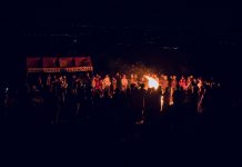 A large group of people gathered around a bonfire at night, featuring a vibrant outdoor atmosphere.