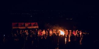 A large group of people gathered around a bonfire at night, featuring a vibrant outdoor atmosphere.