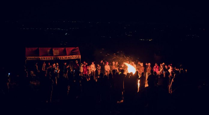 A large group of people gathered around a bonfire at night, featuring a vibrant outdoor atmosphere.