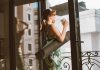 Starbucks and CorePower Yoga Head to Hoboken’s Willow House Woman in workout attire holding a coffee cup, standing on a balcony with a yoga mat, looking outside.