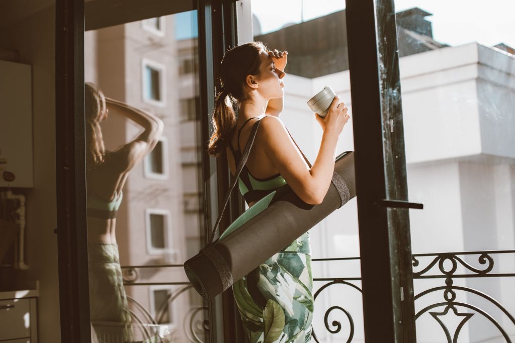 Woman in workout attire holding a coffee cup, standing on a balcony with a yoga mat, looking outside.