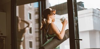 Starbucks and CorePower Yoga Head to Hoboken’s Willow House Woman in workout attire holding a coffee cup, standing on a balcony with a yoga mat, looking outside.