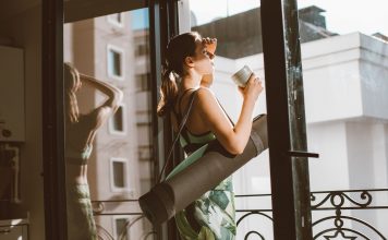 Starbucks and CorePower Yoga Head to Hoboken’s Willow House Woman in workout attire holding a coffee cup, standing on a balcony with a yoga mat, looking outside.