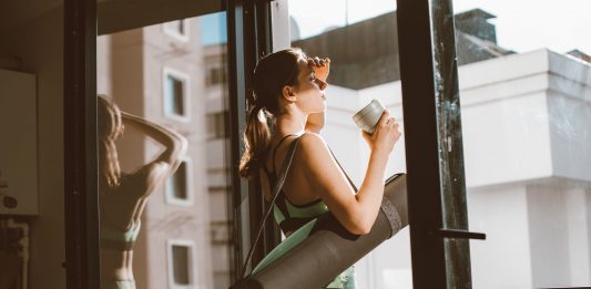 Starbucks and CorePower Yoga Head to Hoboken’s Willow House Woman in workout attire holding a coffee cup, standing on a balcony with a yoga mat, looking outside.