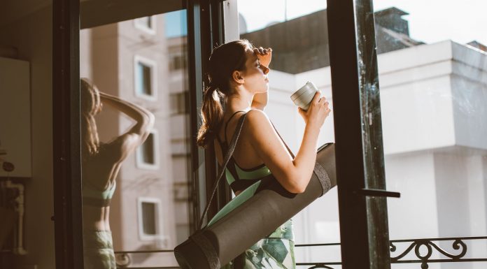 Woman in workout attire holding a coffee cup, standing on a balcony with a yoga mat, looking outside.