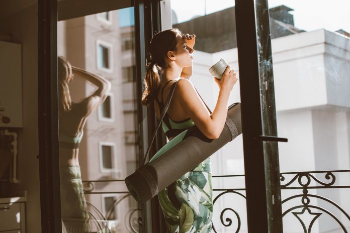 starbucks-and-corepower-yoga-head-to-hoboken-s-wil Woman in workout attire holding a coffee cup, standing on a balcony with a yoga mat, looking outside.