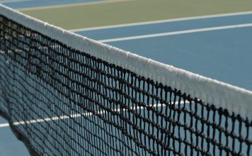 Where to Play Tennis in Jersey City + Hoboken for Free Detailed perspective of a tennis net casting shadows on a vibrant blue court under sunlight.