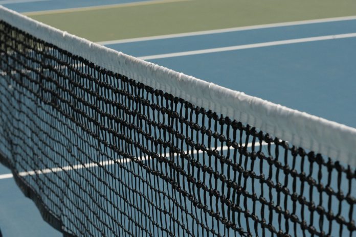where-to-play-tennis-in-jersey-city-hoboken-for-fr Detailed perspective of a tennis net casting shadows on a vibrant blue court under sunlight.