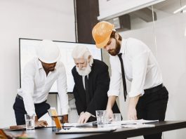 Group of architects discussing construction blueprints wearing hardhats in a modern office.