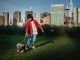 Ethnic child with Yorkshire Terrier on leash preparing to run on meadow against urban skyscrapers