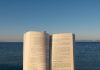 BREAKING: Hoboken Girl Goes on Summer Break Until July 13 An open book is held by the ocean under the calming sunset sky.