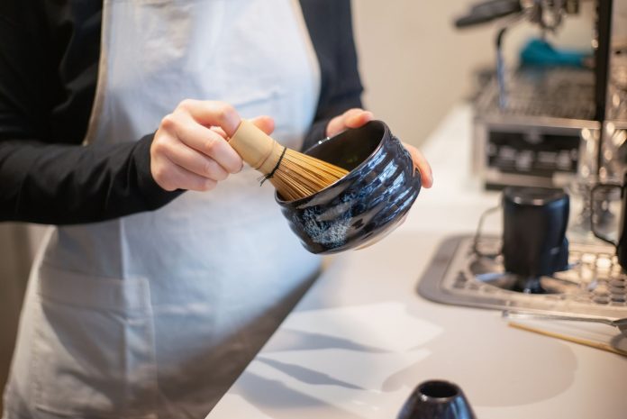 Close-up of a person preparing matcha tea using a bamboo whisk and bowl indoors.