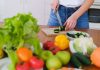Woman slicing cucumbers surrounded by fresh produce in a bright kitchen setting.