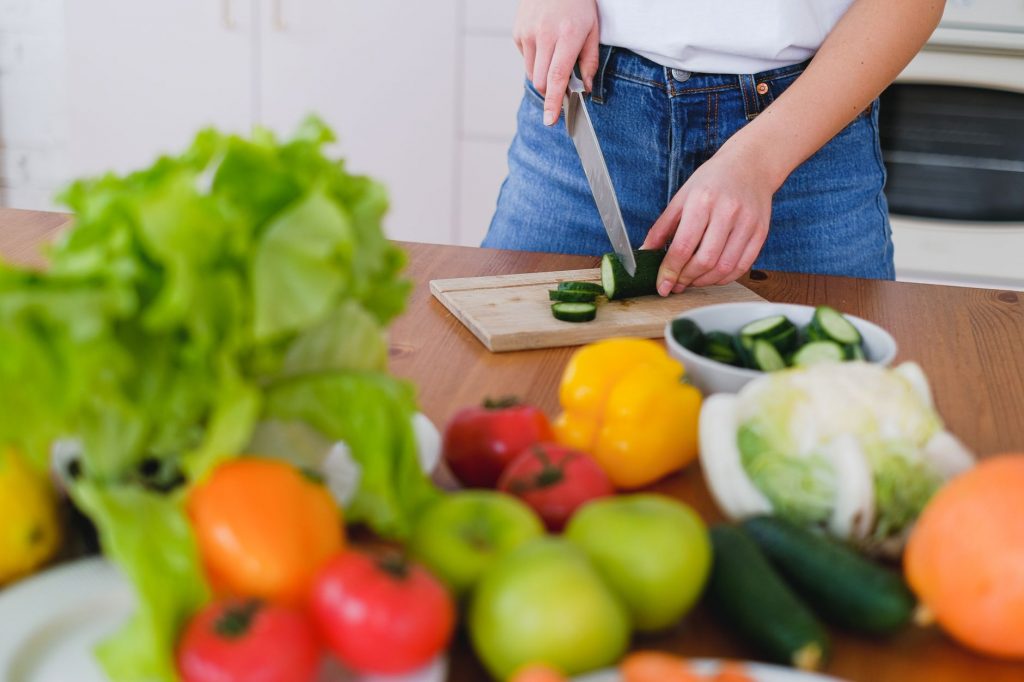 Woman slicing cucumbers surrounded by fresh produce in a bright kitchen setting.