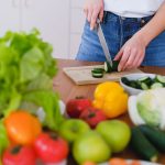 Woman slicing cucumbers surrounded by fresh produce in a bright kitchen setting.