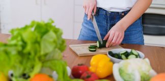 Woman slicing cucumbers surrounded by fresh produce in a bright kitchen setting.