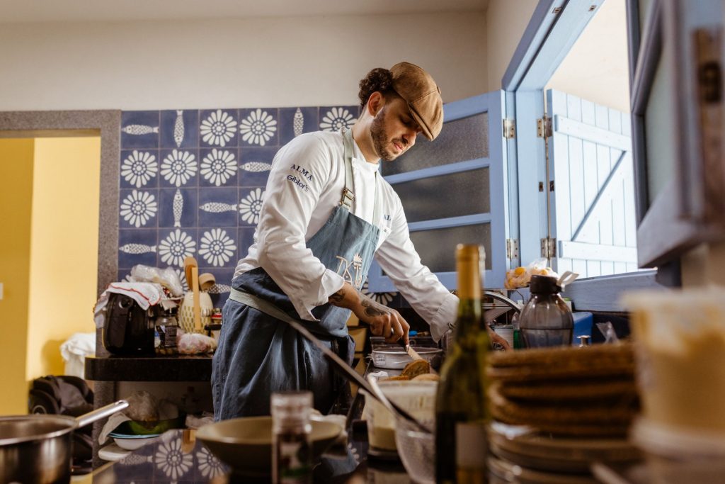 A chef in a rustic kitchen cooks a gourmet meal with an open door filling the room with natural light.