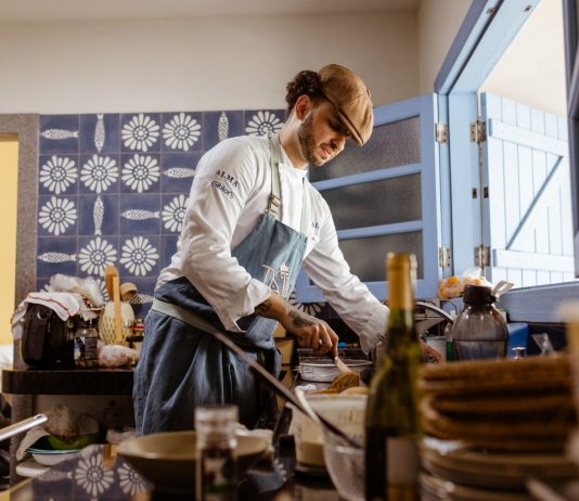 A chef in a rustic kitchen cooks a gourmet meal with an open door filling the room with natural light.
