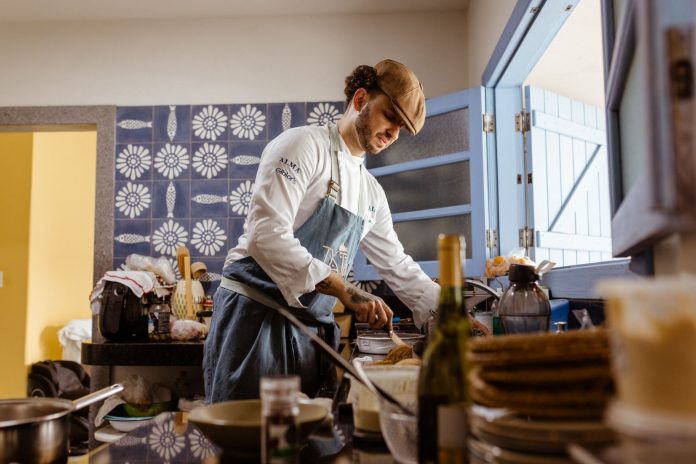 A chef in a rustic kitchen cooks a gourmet meal with an open door filling the room with natural light.