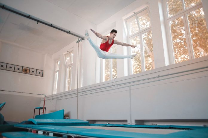 Young sportsman in sportswear doing split in air while jumping on trampoline in gym