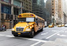 A yellow school bus navigates a busy New York City street amidst tall buildings and taxis.