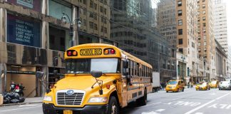 A yellow school bus navigates a busy New York City street amidst tall buildings and taxis.