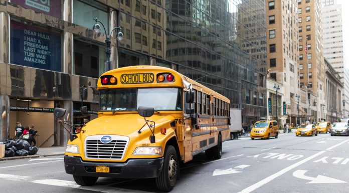 A yellow school bus navigates a busy New York City street amidst tall buildings and taxis.