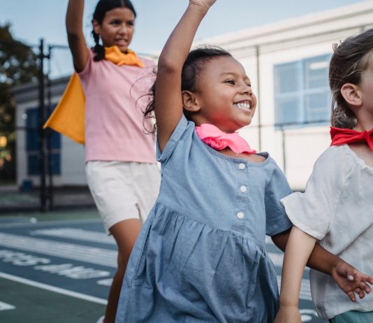 Three kids enjoying superhero play on a sunny outdoor court.