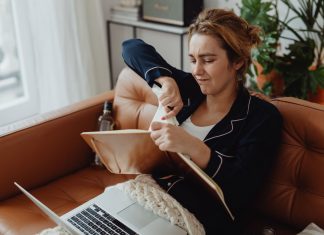 Woman in pajamas tearing paper on a sofa, expressing frustration. Indoors setting with laptop nearby.