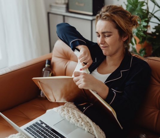 Woman in pajamas tearing paper on a sofa, expressing frustration. Indoors setting with laptop nearby.