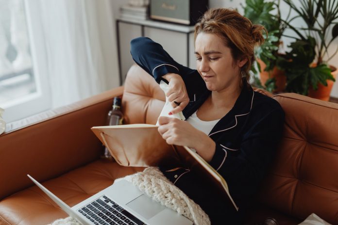 jersey-city-resident-blasts-15-5-tax-hike-calls-bu Woman in pajamas tearing paper on a sofa, expressing frustration. Indoors setting with laptop nearby.