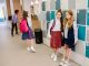 Students in school uniforms stand by lockers in a bright corridor.