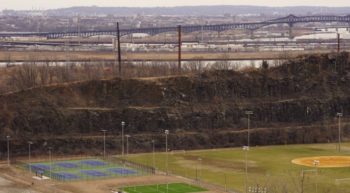Aerial view of multi-sport fields in urban park with city skyline and bridge in background.
