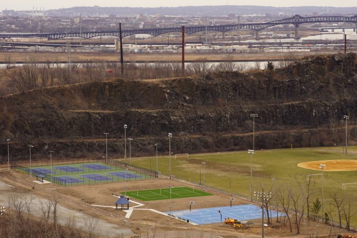lo-que-los-locales-aman-de-vivir-en-elizabeth Aerial view of multi-sport fields in urban park with city skyline and bridge in background.