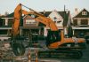 Excavator working in a suburban neighborhood construction site, with residential houses in the background.